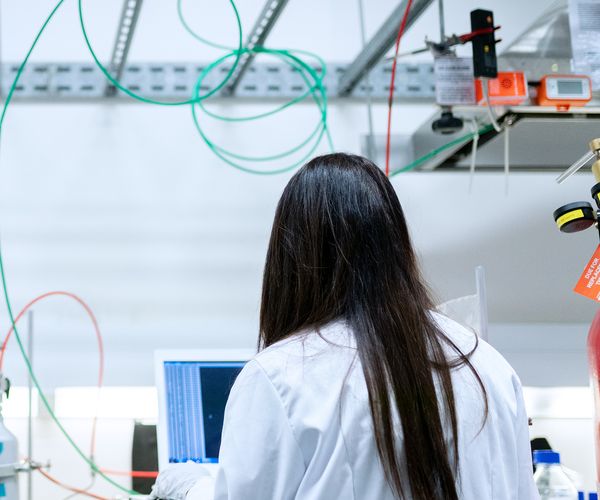 An individual with long hair stands at a computer in a laboratory setting.