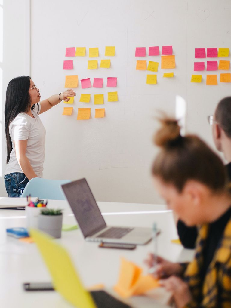 A female presenting person stands at a white board pointing at a bunch of post it notes.