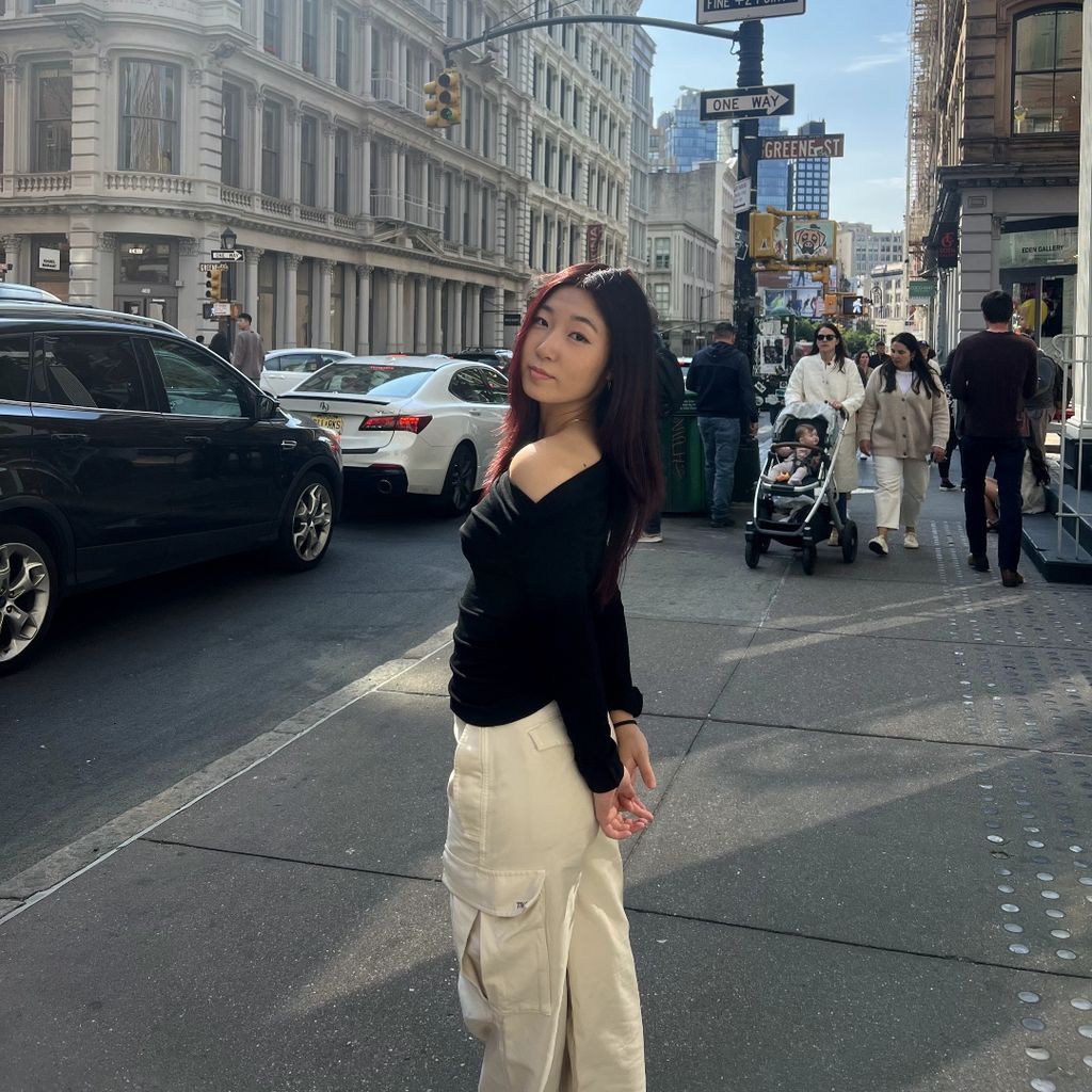 BA Music student Grace Yuk posing for a photo on a busy street in front of Greene St. Grace is wearing beige cargo pants and a black long sleeve shirt with her shoulder out.