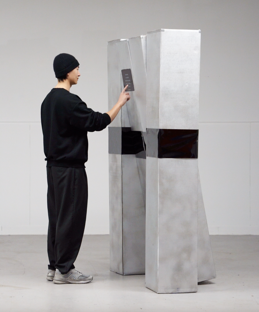 MFA Graphic Design student Wenbin Huang is seen touching a Robot Interaction Design project, titled Time Space Love. Wenbin is wearing a black beanie hat, black sweat shirt, black joggers, and New Balance sneakers. The installation is tall and gray.