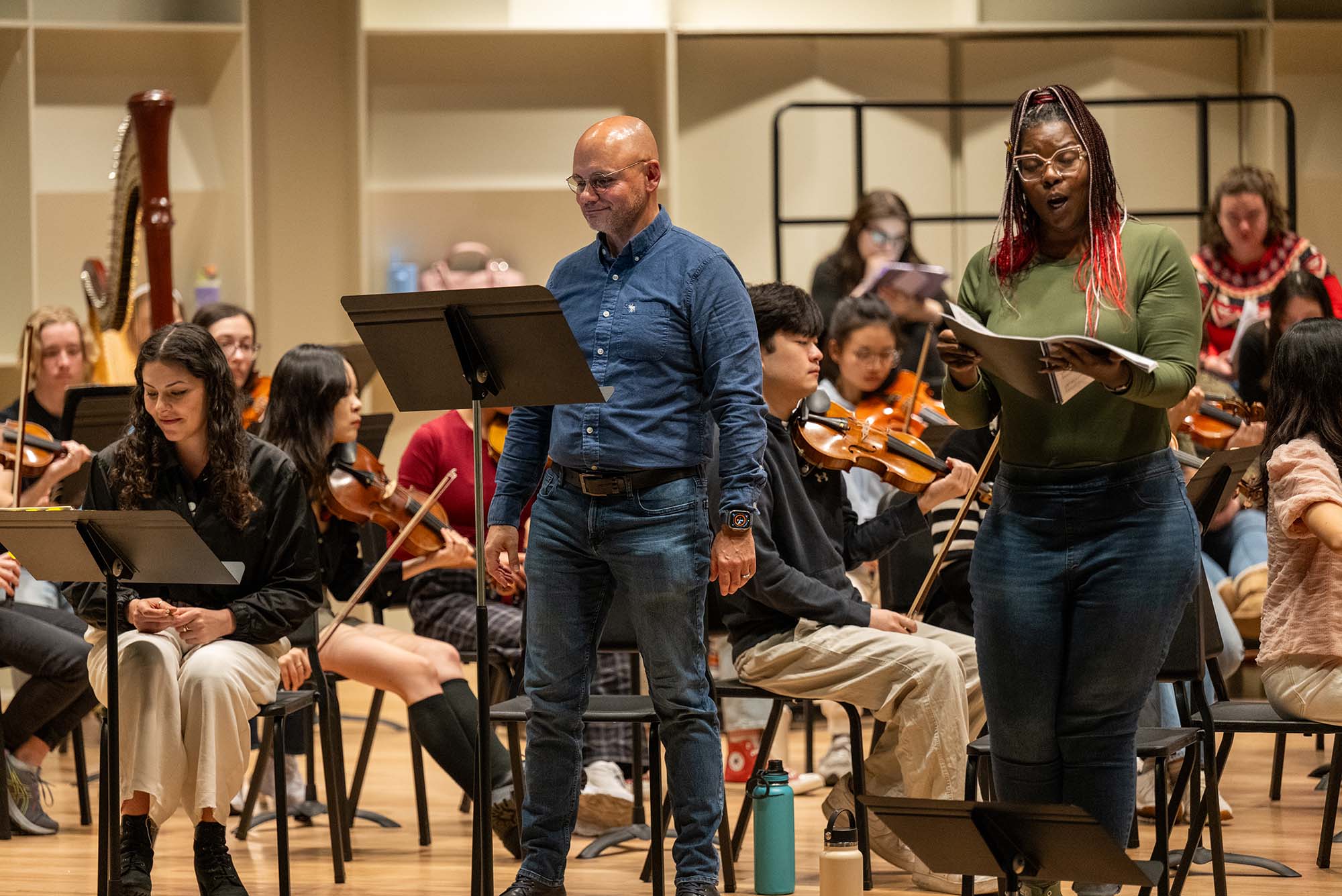 A rehearsal scene with musicians and vocalists. In the foreground, Boston University assistant professor of music, voice, David Guzmán, wears glasses and a blue shirt and is standing near two singers. The singer to his right (stage left) is mezzo-soprano student Juliette Kaoudji who is seated and smiling. The other singer, standing on the opposite side of David, is soprano Michelle Johnson, who is singing while holding sheet music. In the background, an orchestra with string players and a harpist rehearses.