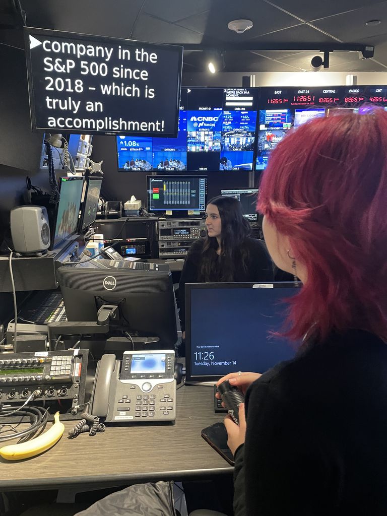 A person with vibrant pink hair is seen from behind, holding a communication device while observing a control room filled with monitors, computers, and broadcasting equipment. Another individual with long dark hair is seated at a workstation, focused on the screens. A large display in the room shows text about a company being in the S&P 500 since 2018. The environment is dimly lit, illuminated primarily by the screens and equipment
