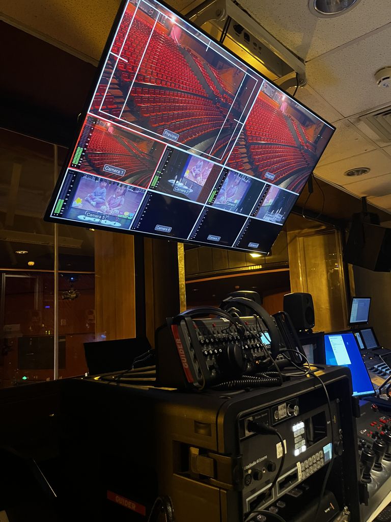 A large monitor displays a multi-camera feed in a control room, showing various angles of an empty theater with rows of red seats and some additional video feeds showing children on stage. Below the screen, professional broadcasting and audio equipment, including a communication device labeled 'GUIDER,' is stacked on a black case. The dimly lit environment is filled with screens, buttons, and controls, reflecting a professional production setup.
