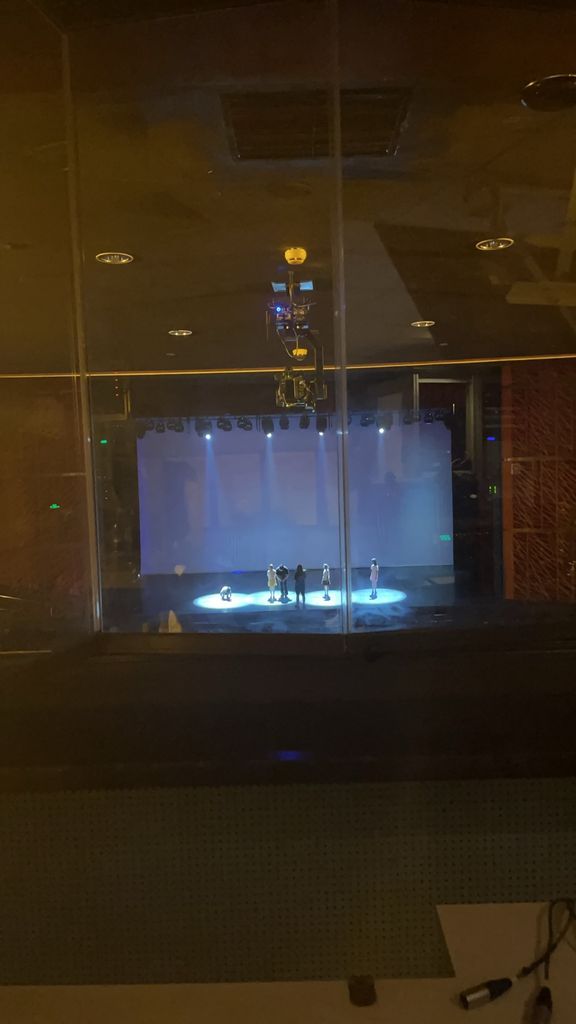 A view from a control booth looking out onto a theater stage where a small group of people is gathered under bright spotlights. The stage is illuminated with cool-toned lighting, while the foreground is darker, showing a reflection of the booth’s ceiling and equipment on the glass window. A mounted camera is visible hanging from the ceiling, capturing the scene below.