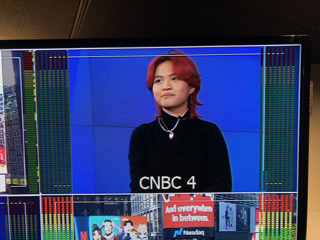 A close-up of a television broadcast monitor displaying a person – Miya Menscher - with short, red hair wearing a black turtleneck and silver necklaces. There is text 'CNBC 4' visible on the screen. The broadcast monitor includes colorful audio level indicators and a split-screen effect showing part of a Nasdaq billboard with the text 'and everywhere in between.’