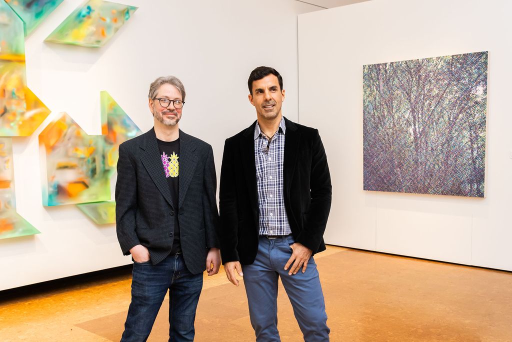 Colleagues and friends Michael Zachary (left) and Juan José Barboza-Gubo standing in BU's Faye G., Jo, and James Stone Gallery for their joint exhibition, Trasluz / Translucent. The man on the left wears glasses, a blazer, jeans, and a T-shirt with colorful pineapples. The man on the right is dressed in a plaid shirt, blue pants, and a dark velvet blazer. Behind them, on the left, is a three-dimensional geometric installation with bright, translucent colors. On the right, a square canvas displays an intricate, abstract pattern resembling a forest scene. The gallery has a clean, modern interior with bright lighting and a polished wooden floor.