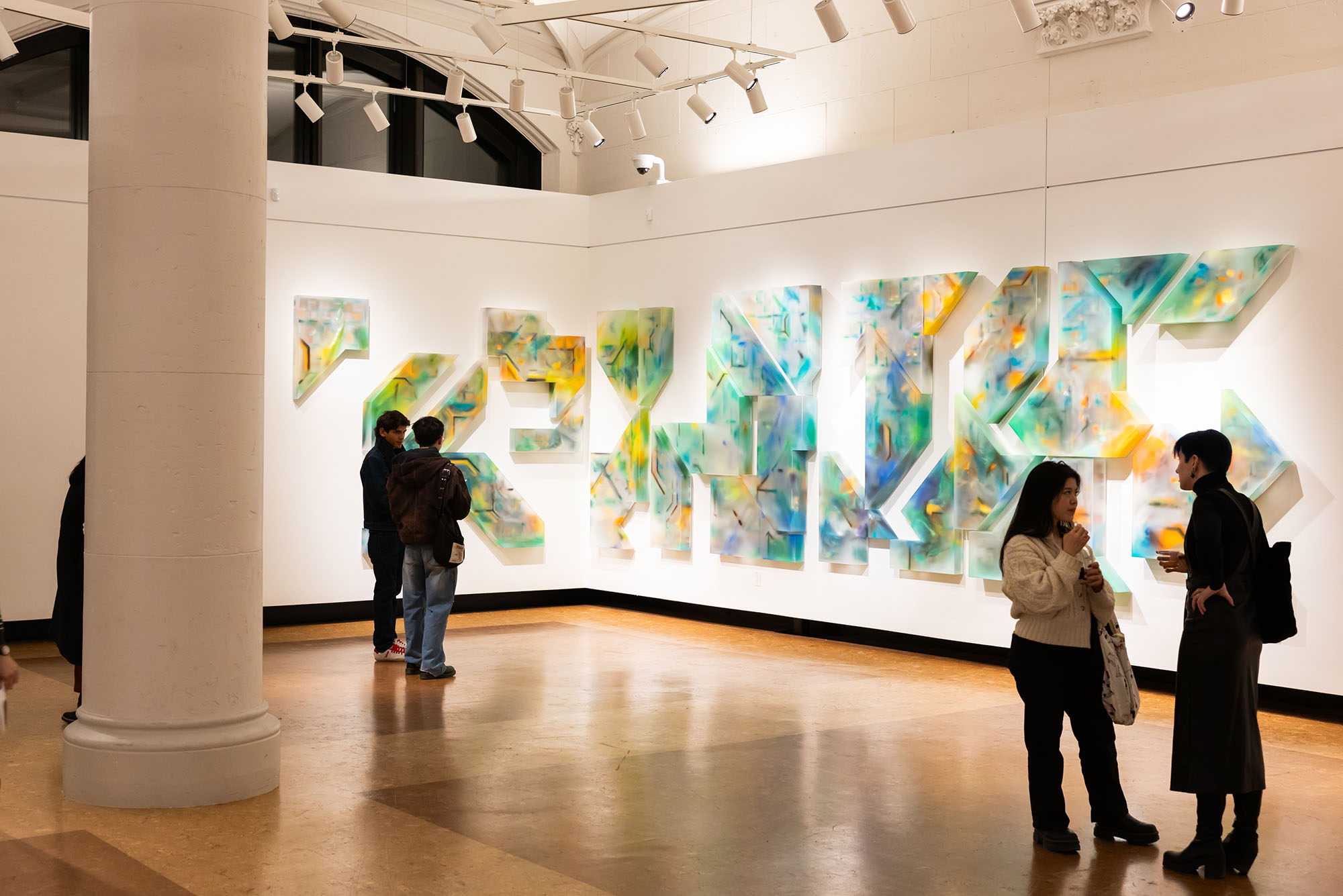 BU Art Galleries' Faye G., Jo, and James Stone Gallery latest exhibition, Trasluz / Translucent. The image shows the gallery with a vibrant, abstract installation on the wall featuring three-dimensional geometric shapes with colorful, translucent patterns in shades of blue, green, yellow, and orange. The pieces are arranged in a way that creates an interconnected, dynamic composition. Four people are engaged in conversation in front of the artwork, creating a lively atmosphere. The gallery is brightly lit with ceiling spotlights, and the polished wooden floor reflects the light, enhancing the spacious and modern setting. A large white pillar partially frames the left side of the image.