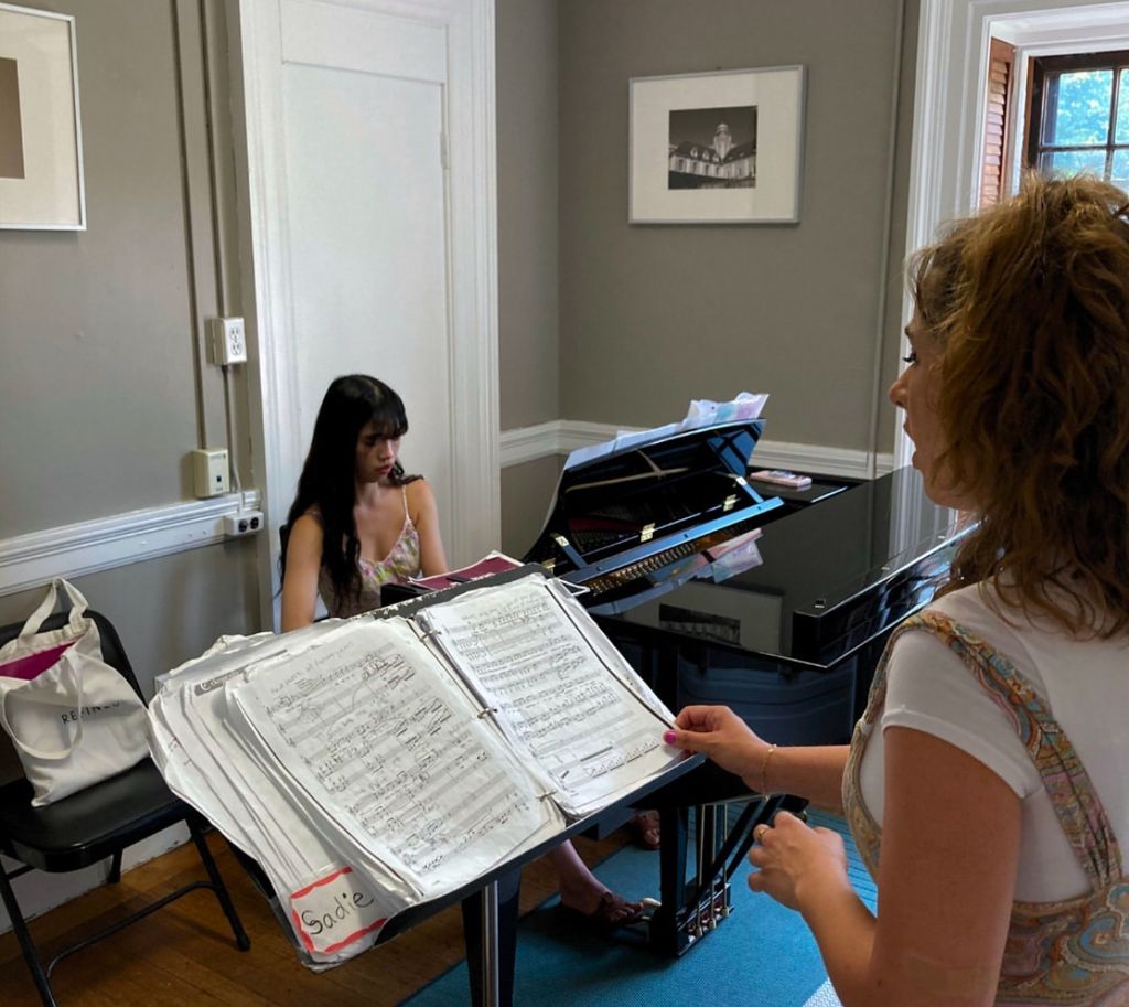 Two women are in a music rehearsal room. One is seated at a grand piano, focused on playing from sheet music, her long dark hair cascading over her shoulders. The other stands beside a music stand labeled "Sadie," singing and gesturing expressively. The room is softly lit with framed pictures on the neutral-colored walls and a window with wooden shutters in the background, creating an intimate, creative atmosphere.