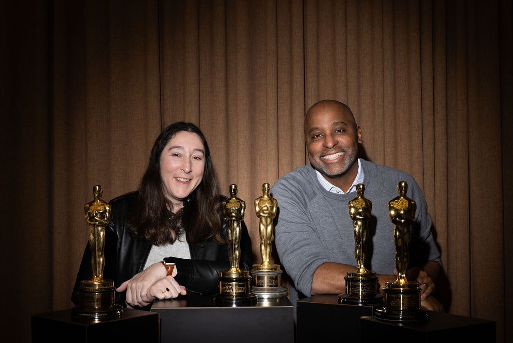 Betsy Walters (COM’20, GRS’25) and Harvey Young, dean of the College of Fine Arts smiling and posing with five Oscar statuettes displayed on black pedestals in front of them. They stand against a backdrop of brown curtains. Walters (left) has long dark hair and is wearing a black leather jacket, while Dean Young on the right is wearing a gray sweater.