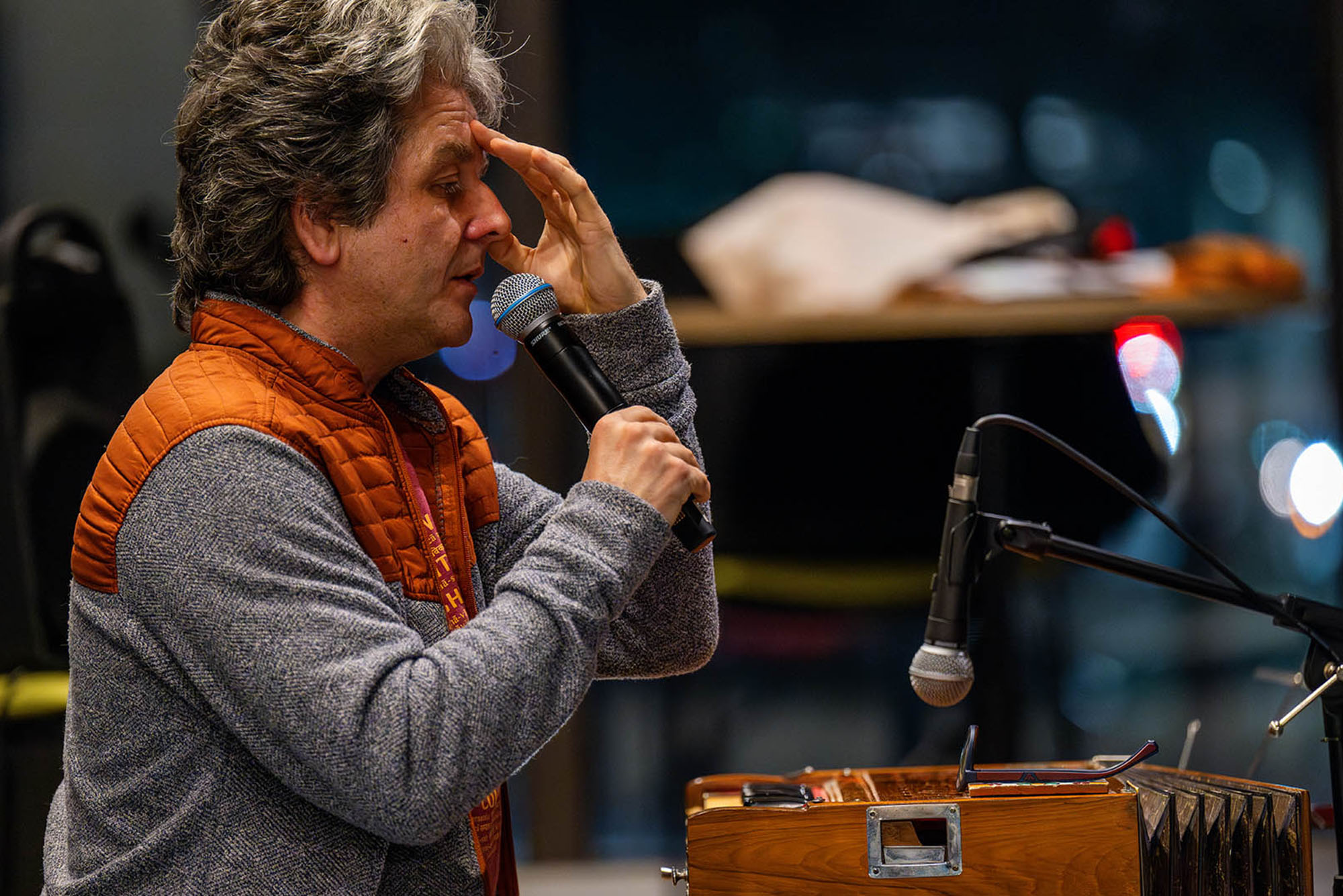 CFA’s Igor Iwanek seen with wavy gray hair and light skin holds a microphone while speaking at one of the breath workshops he leads, with his other hand resting on his forehead. He is wearing a gray sweater with an orange quilted vest. In front of him is a wooden harmonium with a microphone positioned nearby. The background is blurred, with warm indoor lighting and hints of a reflective surface, possibly a window or glass wall, with city lights visible outside.