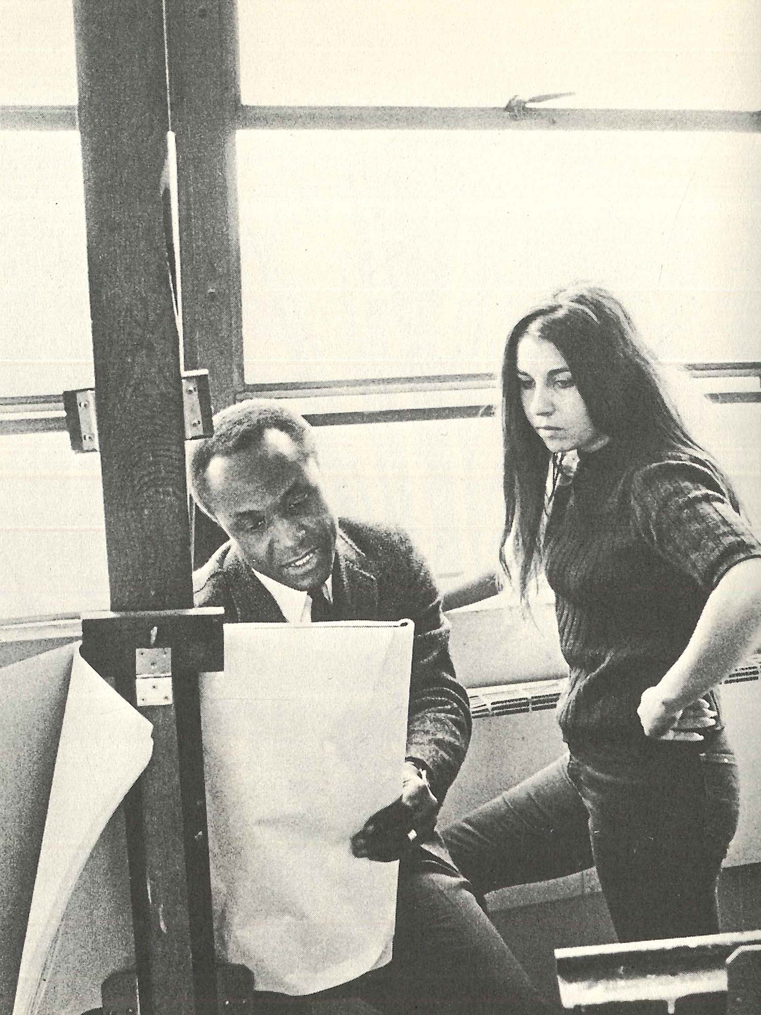 A black-and-white photo of the late artist and Boston University professor John Wilson and a student engaged in a discussion. Wilson, seated on the left, holds a large sheet of paper on an easel, pointing and speaking animatedly. The student, standing with one hand on her hip, listens attentively, her long hair falling over her shoulder. They are in a bright studio space with large windows in the background, casting soft light over the scene, emphasizing the collaborative and educational atmosphere.