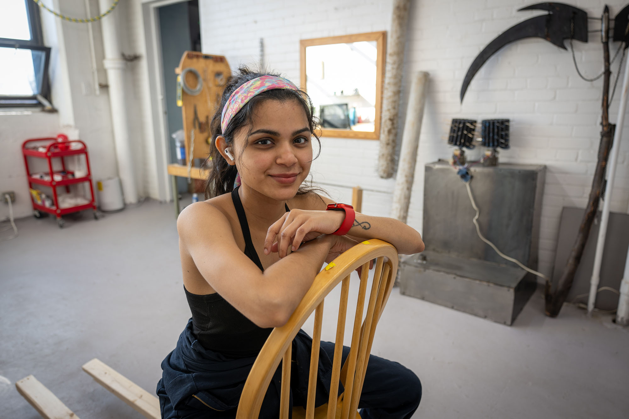 Maithili Rajput, a Boston University graduate sculpture student, with dark hair tied back with a colorful headband sits backward on a wooden chair in an artist’s studio. She wears a black tank top, navy work pants, a red watch, and wireless earbuds, smiling gently at the camera. The background features a white brick wall adorned with sculptural tools and materials, including a large black metal scythe, wooden logs with spikes, and a workstation with various equipment. Sunlight streams in from a window, casting a warm glow on the creative workspace.