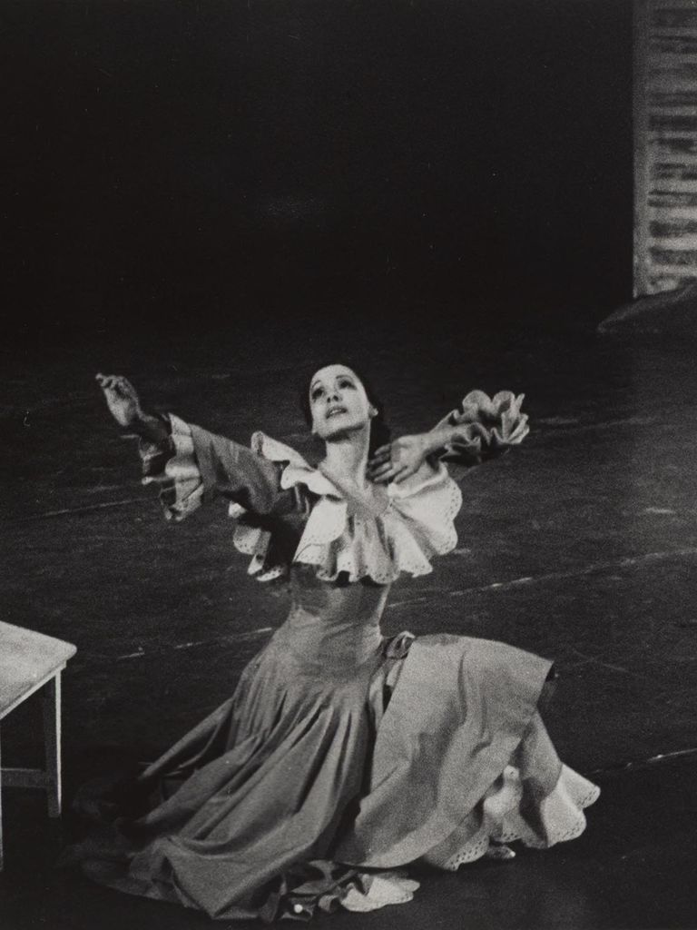 A black-and-white photograph captures a dramatic moment in a dance performance. The late Pearl Lang, an American dancer and choreographer, is in a flowing dress with ruffled sleeves, kneeling on stage, her arms extended outward and her head tilted back with an expressive gaze. The dark stage background contrasts with her lighter costume, emphasizing the motion and emotion in her performance. A small wooden bench is visible to the side, adding context to the theatrical setting.