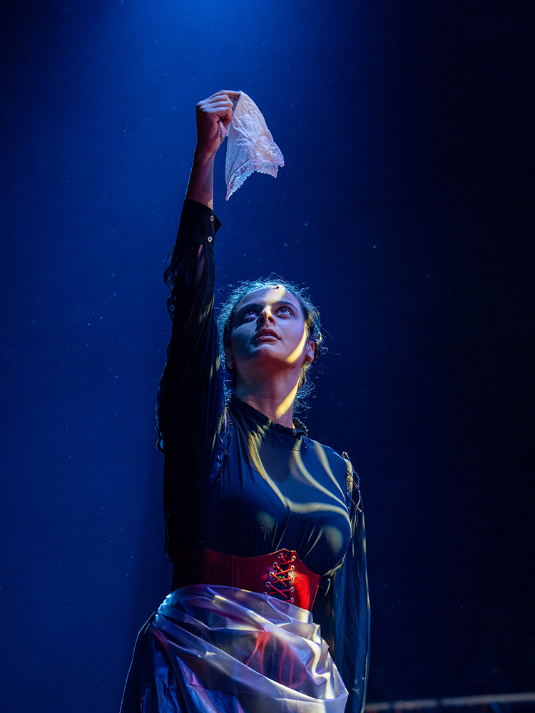 An actor holding a handkerchief high in the air on stage against a dark backdrop