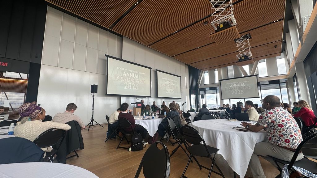A wide-angle view of the 波士顿大学 Center on Forced Displacement Annual Conference 2025. Attendees sit at round tables in a high-ceilinged room with floor-to-ceiling windows. Multiple large screens display the conference title at the front of the room.