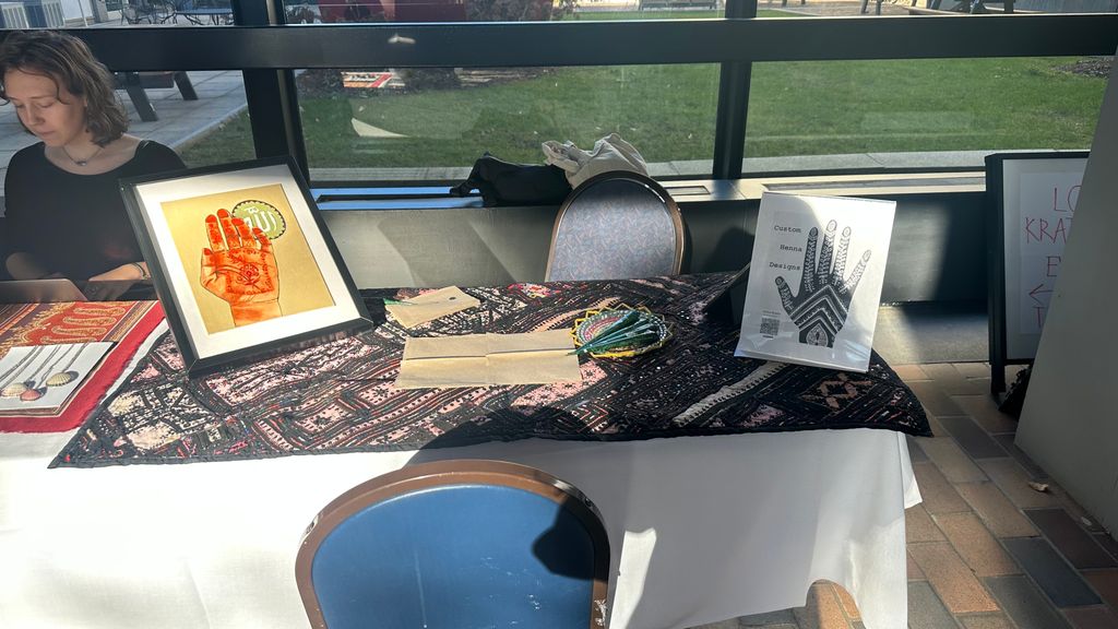 A tabletop display set up for custom henna designs. The table is covered with a richly patterned cloth and features framed artwork of a henna-covered hand, a small sign labeled “Custom Henna Designs,” beaded jewelry, and informational flyers. A woman is seated behind the table, partially out of frame.
