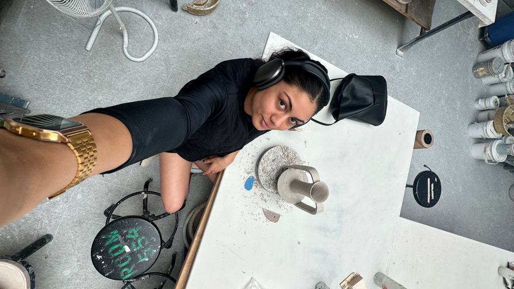 A selfie taken by an artist in a ceramics studio, looking up at the camera while seated at a white table. She wears black clothing and over-ear headphones, with pottery tools and a sculptural ceramic pitcher in progress in front of her. The background shows stools, buckets, and studio equipment.