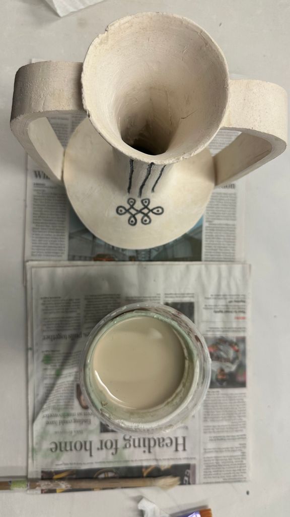 Overhead shot of a finished bisque-fired ceramic pitcher with two handles, placed on newspaper. The pitcher features hand-drawn black designs and sits next to a jar of creamy glaze, ready for application.