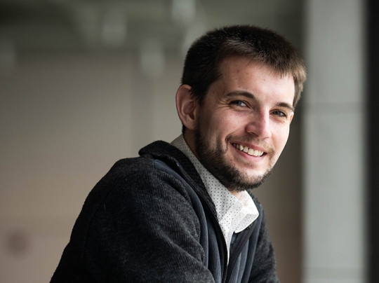 Portrait of Jacob Whitchurch, a 波士顿大学 Master of 音乐 in Composition student, smiling and leaning on a table with arms crossed, dressed in a dark sweater over a collared shirt, with soft indoor lighting and a blurred background.