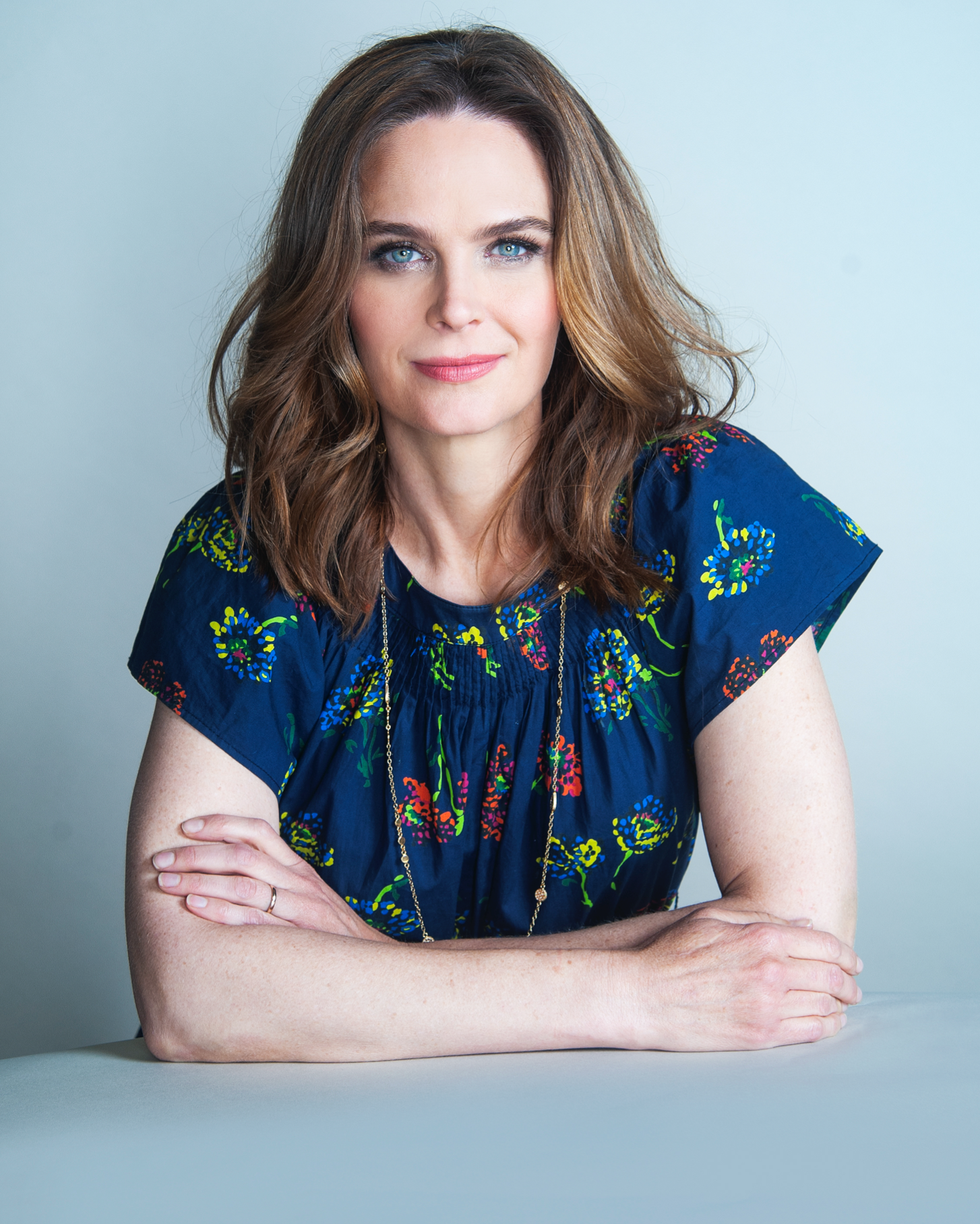 Actor 达斯夏奈 with wavy brown hair and blue eyes sits at a table with her arms crossed, wearing a navy blue top with a colorful floral pattern and a long necklace, looking confidently at the camera against a light gray background.
