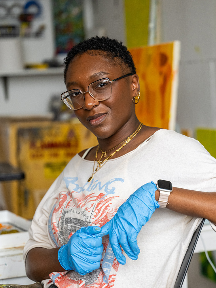 波士顿大学 MFA Painting student Raquel Philippe sits in a studio space wearing a graphic T-shirt, glasses, and blue disposable gloves. She looks confidently at the camera, with one arm crossed and the other adjusting a glove. She wears layered gold necklaces, hoop earrings, and an Apple Watch. Behind her are colorful abstract artworks and studio materials.