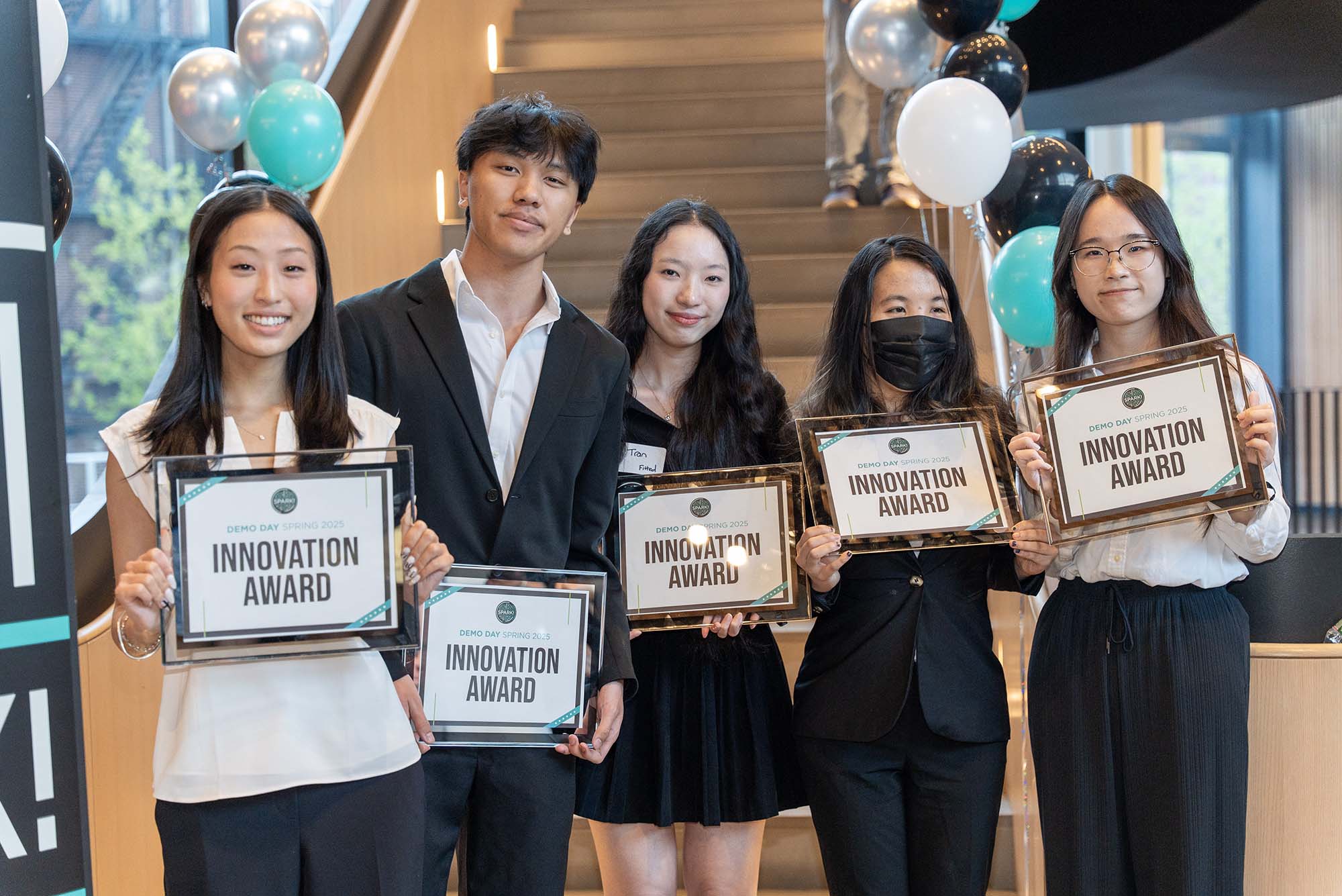 Five students stand smiling and holding framed certificates labeled “Innovation Award” at BU火花! 演示日 Spring 2025. They are dressed in business or semi-formal attire, and balloons decorate the staircase behind them.