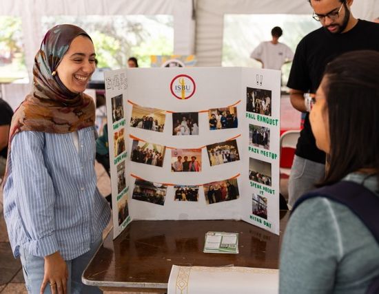 A member of ISBU speaks with another student and shows photos of previous events on a posterboard