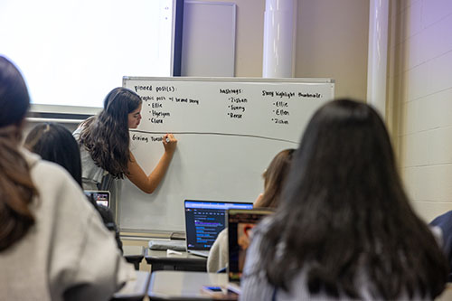 Woman writing on a white board in front of a group of peers.