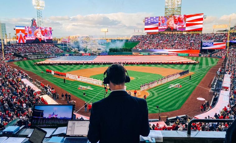Back of man wearing headset and is facing a baseball field during the national anthem with a large American flag.