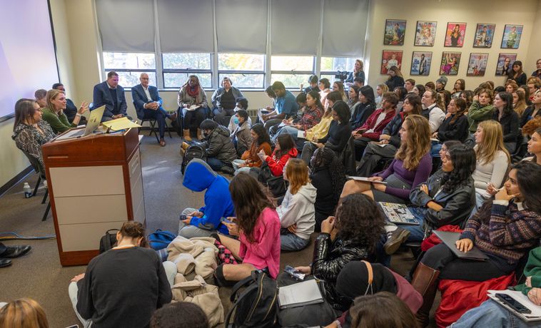 A large group of students listening to a panel of speakers in a classroom.