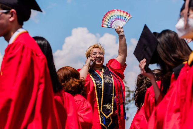 Photo of a tan person with short, bronze bleach hair smiling as they holds a cellphone to their ear and waves a rainbow fan that reads "#proud". They wear a black says that reads "Alianza Latina". Grads surround her wearing similar gowns and caps.