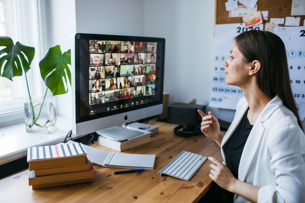 A woman sits at a desktop computer. On the screen is a video meeting with a large grid of many participants' video feeds.