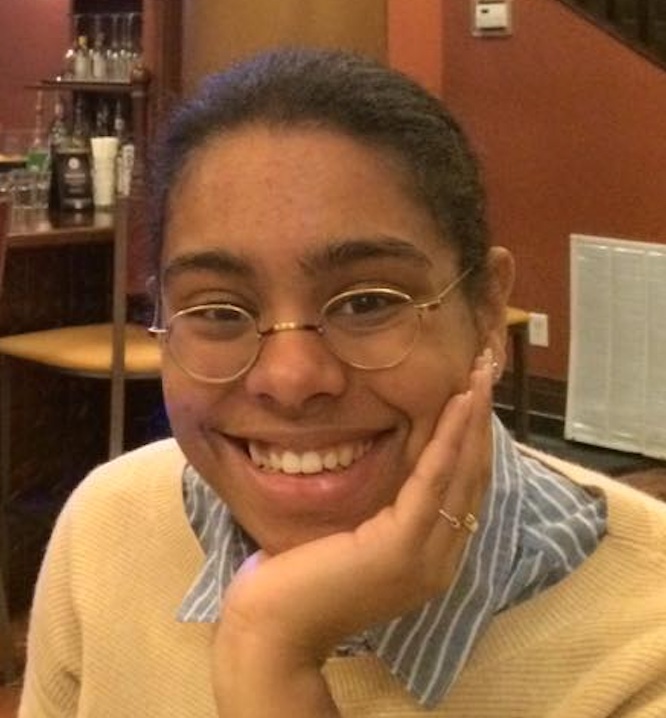 Close up headshot of a woman with dark hair pulled back and wearing glasses. Has a blue and white striped collared shirt under a yellow sweater. hand resting in her chin.