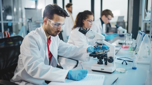 Students working in a laboratory are looking at samples under a microscope and writing in a notebook