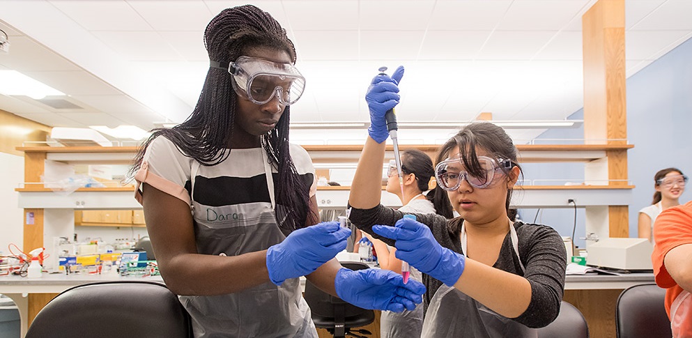 Two women wearing protective gear work together in a lab to fill test tubes
