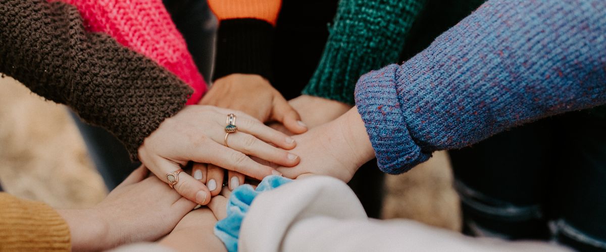 Group of people put their hands into the middle of a circle, each wearing a different colored sweater
