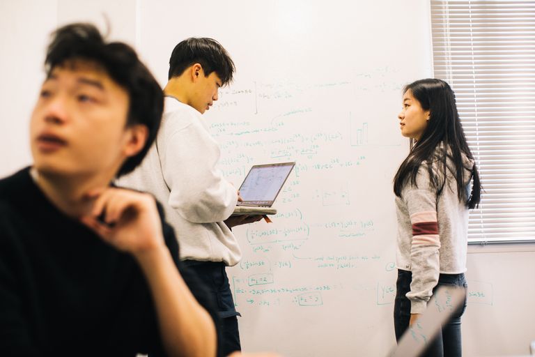 Student holding a laptop while discussing data on a white board with other students