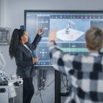 Woman Presenting at Digital Whiteboard to Engineering Class as a student raises their hand