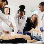 Medical students learning how to conduct CPR as students in white coats surround a classmate practice proper technique on a CPR mannequin