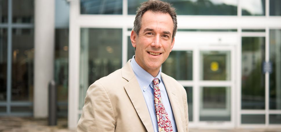 Portrait of Andrew Budson in a tan suit jacket and floral tie in front of a building with a white facade.