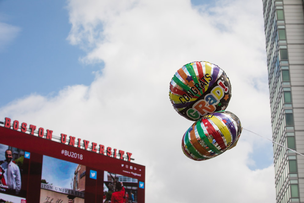 two balloons that say Congrats GRAD! float in the sky next to the Boston University digital sign during commencement