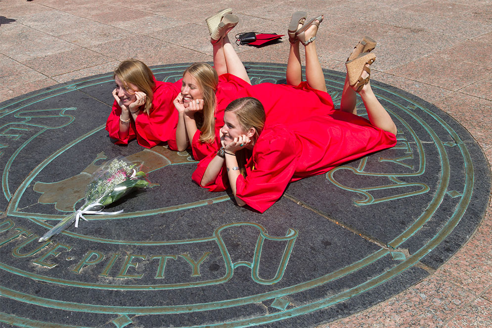3 female Boston University graduates lay on the BU Seal symbol posing for a photo on Marsh Plaza.