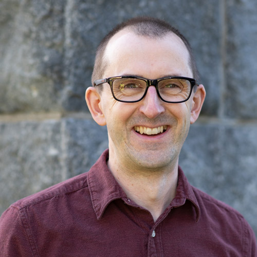 Photo of Andrew Thurston, a white man with black glasses. He smiles and wears a maroon polo shirt.