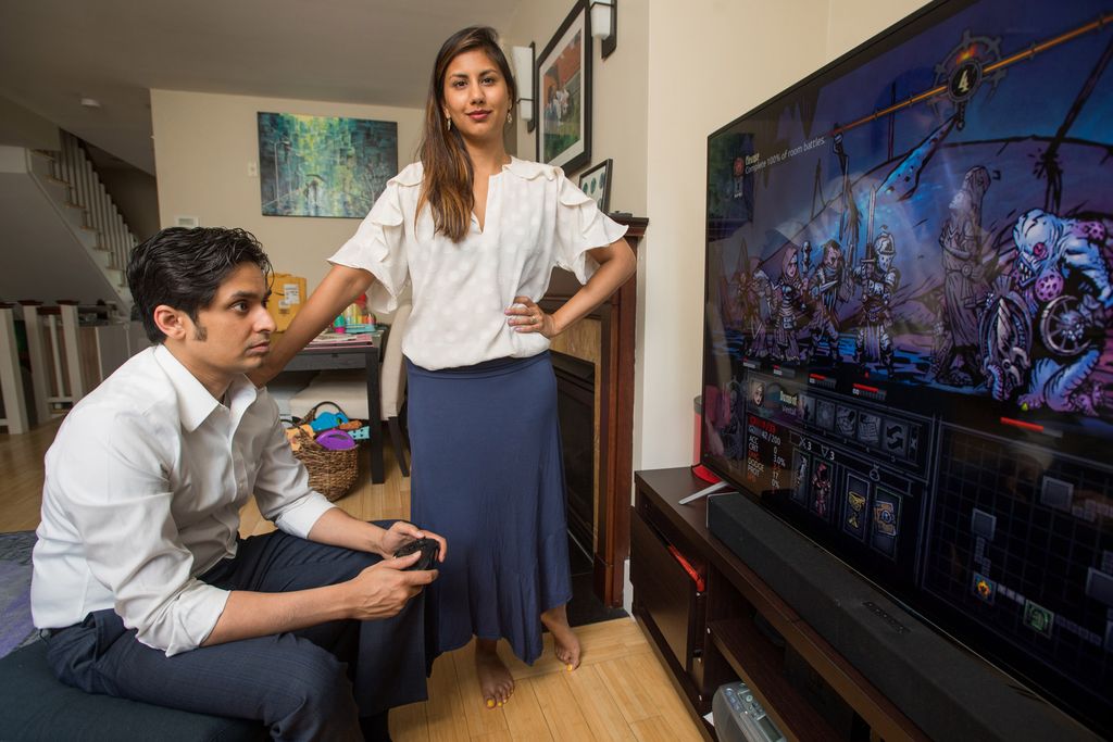 Kruti Kanojia standing next to her husband and cofounder of Healthy Gamer as he plays a video game on a large television.