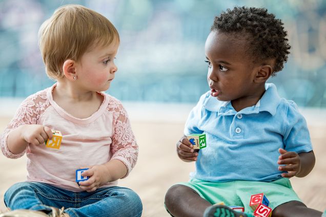 Two toddlers sit and play with alphabet blocks next to each other.