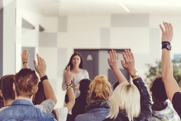 Classroom of students with hands raised, with instructor out of focus in the background.
