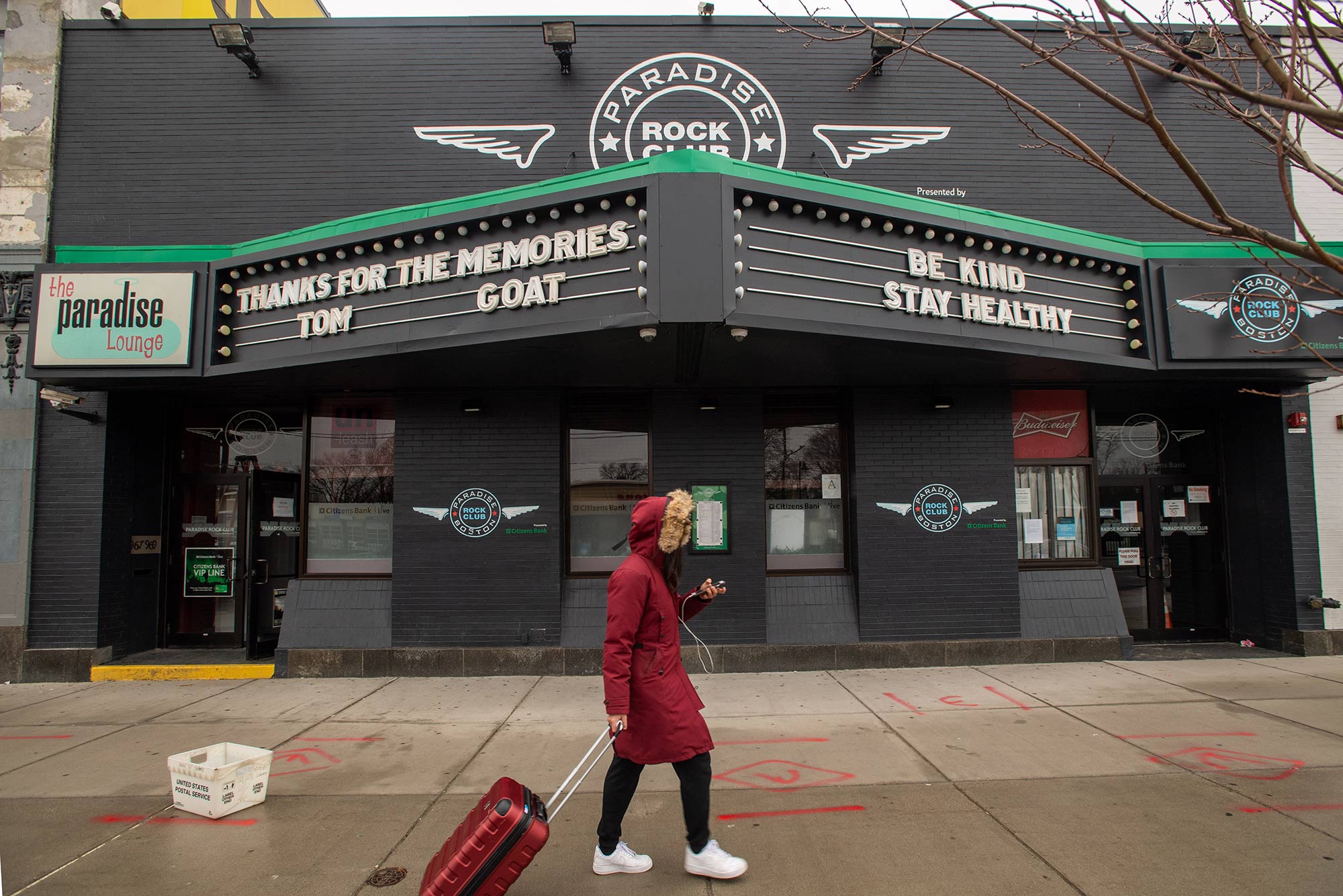 Young person with suitcase walks on the sidewalk. The Paradise Lounge's marquee displays the words "Thanks for the memories, Tom GOAT," and "Be Kind. Stay Healthy". The lounge will be closed for the duration of the coronavirus situation. Photo by Cydney Scott