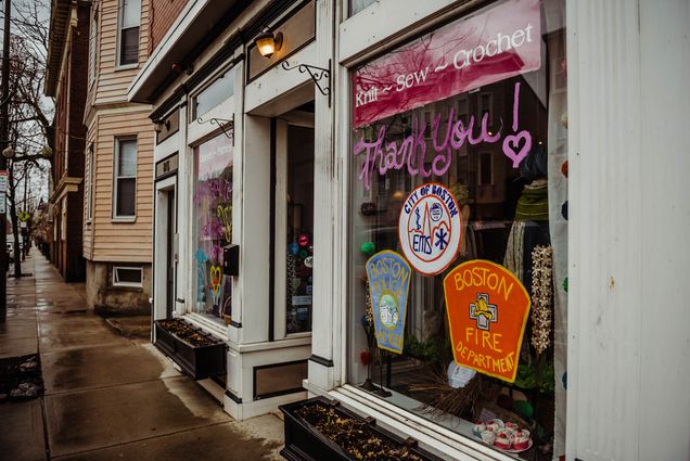 The outside of Stitch House in Dorchester; someone has decorated the front store window to say "Thank you!" in bright pink letters with drawings of the logos from the Boston Fire Department, Boston EMTs, and Boston Police Department underneath. The sidewalk is wet and it's a gloomy day.