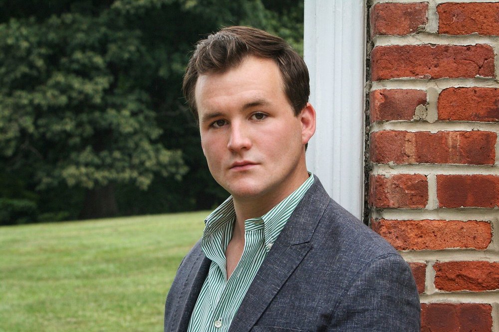 Portrait of Eric Carey in a blazer and button down shirt. Behind him is a brick wall and grass.