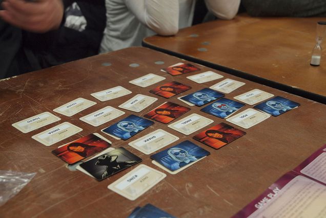 Photo of a game of Codenames being played at the annual board game festival of Finland in Helsinki, Finland. Some cards in the five by five grid show words, while others have blue, red and black illustrations of people.